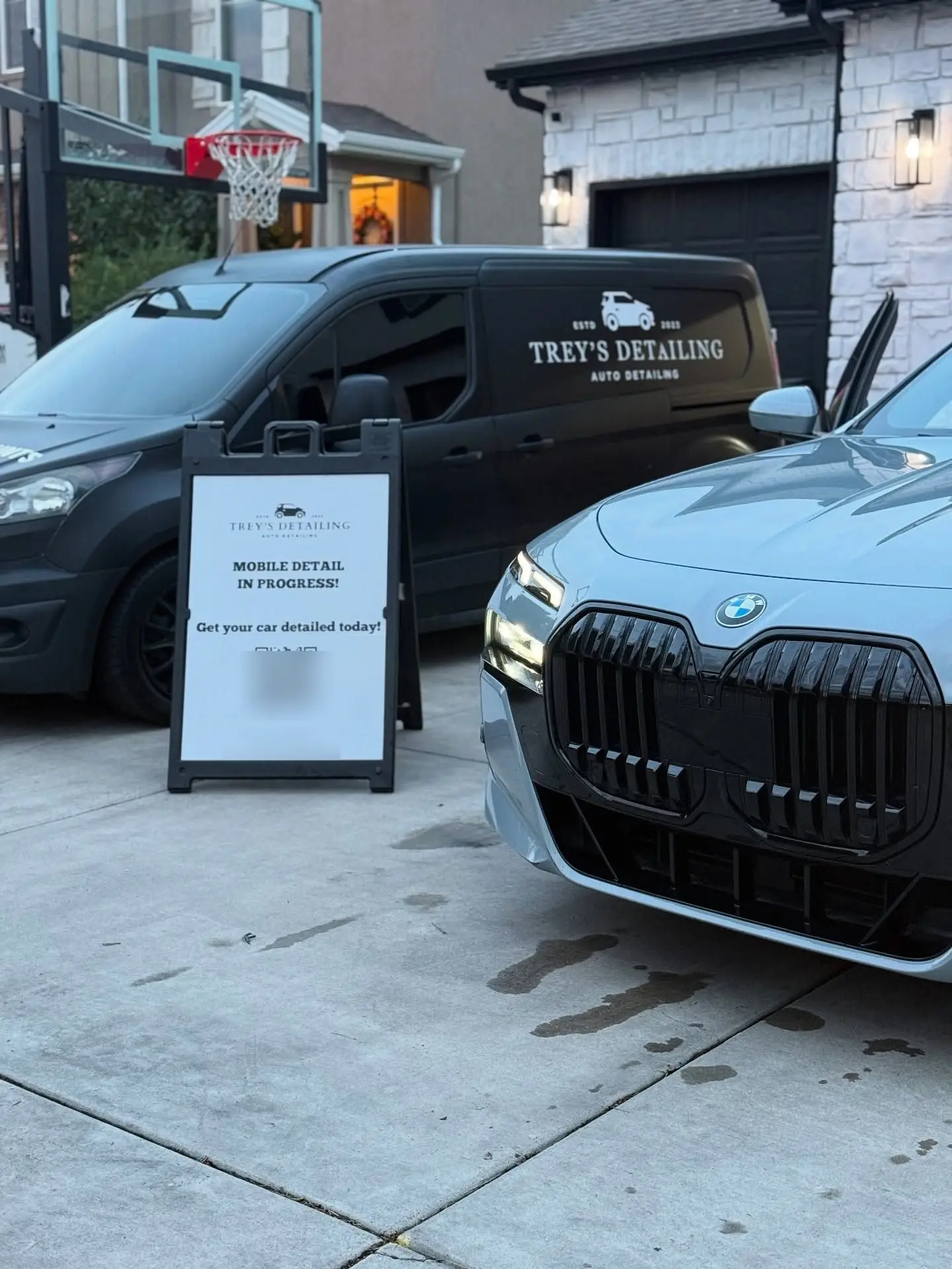 A gray BMW 7 Series on a home driveway beside the Trey's Detailing van and a mobile-detail-in-progress sign