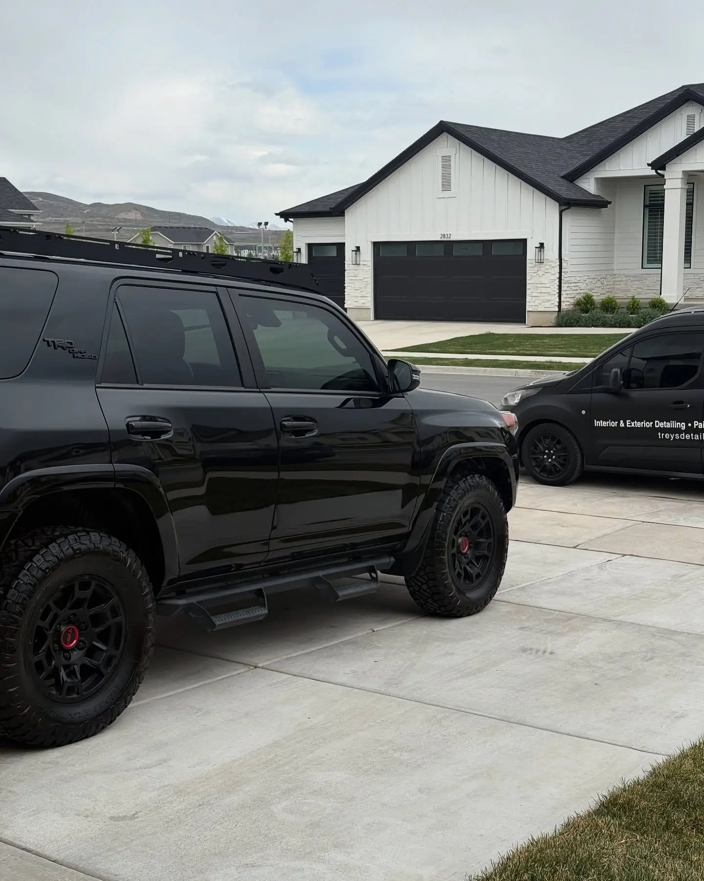 A black Toyota 4Runner parked on a driveway in front of a modern white farmhouse, the detailing van behind it