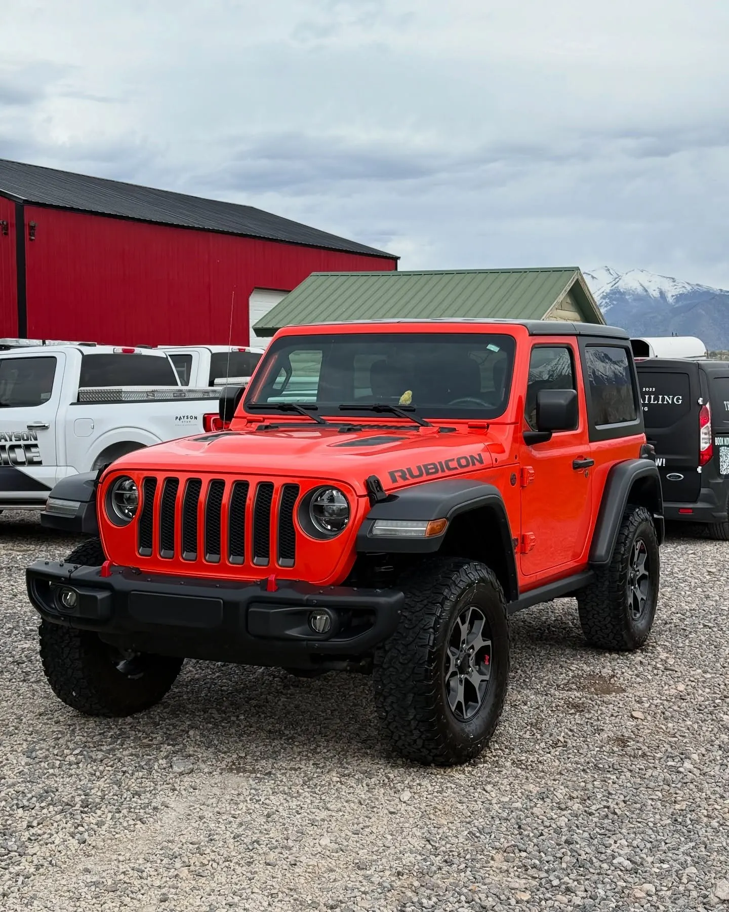 A red Jeep Wrangler Rubicon parked on gravel, trucks behind it